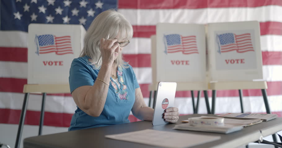 Three quarter angle, medium shot, elderly woman election worker checks voter identification for young female voter using tablet computer at polling station. Flag and voting booths behind