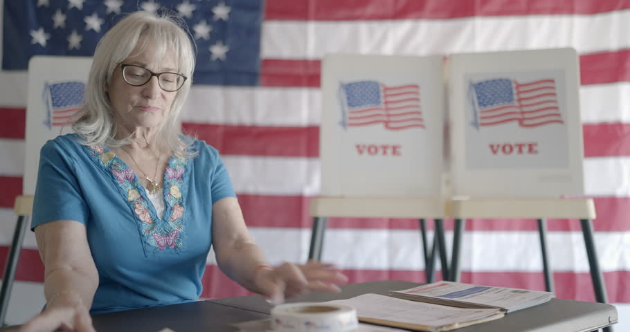 Three quarter angle, medium shot, elderly woman election worker checks over forms at polling station, then smiles to camera as it moves to close up. Flag and voting booths behind