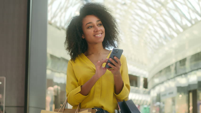 Happy cheerful African American woman standing shopping mall shopaholic fashion discounts black friday buying new things clothes smiling texting chatting smartphone mobile phone gadget connect web sms