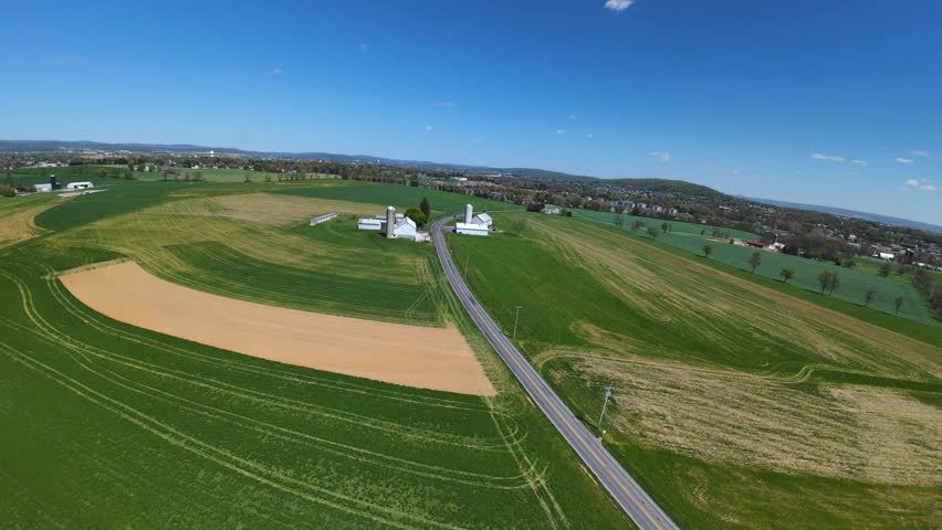 Drone flight over street in rural area with agricultural fields. Sunny day in countryside of USA. Driving car on road. Silo storage at farm. Descending aerial shot.