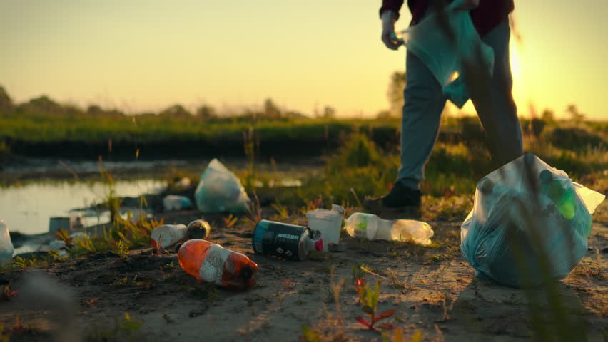 A man wearing gloves collects garbage near a pond. Plastic bottles and other waste go into a trash bag, showing concern for a clean planet. Recycling of disposable tableware, sorting and disposal