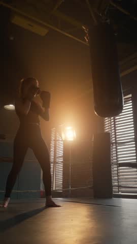 Woman striking punching bag by hands in gym, silhouette of active sportswoman, vertical video. Release of aggression and psychological tension in fitness club, do sport for stay healthy and keep fit