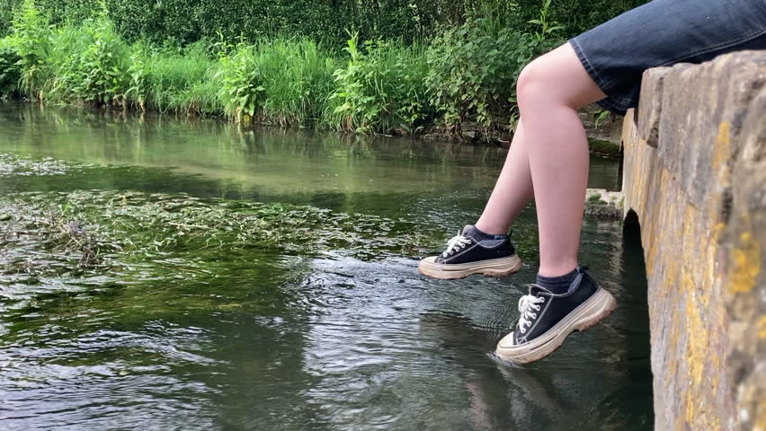 Woman dangling legs over small bridge in Cotswolds with river running under.