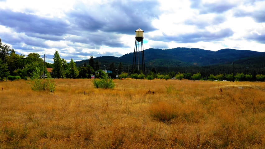 Aerial Forward Ascending Shot Of Tall Water Tower On Green Landscape By Rippled River Under Cloudy Sky - Coeur d