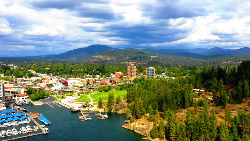 Aerial Panning Scenic View Of Buildings On Green Landscape By Mountains, Drone Flying Over Rippled River During Sunny Day - Coeur d