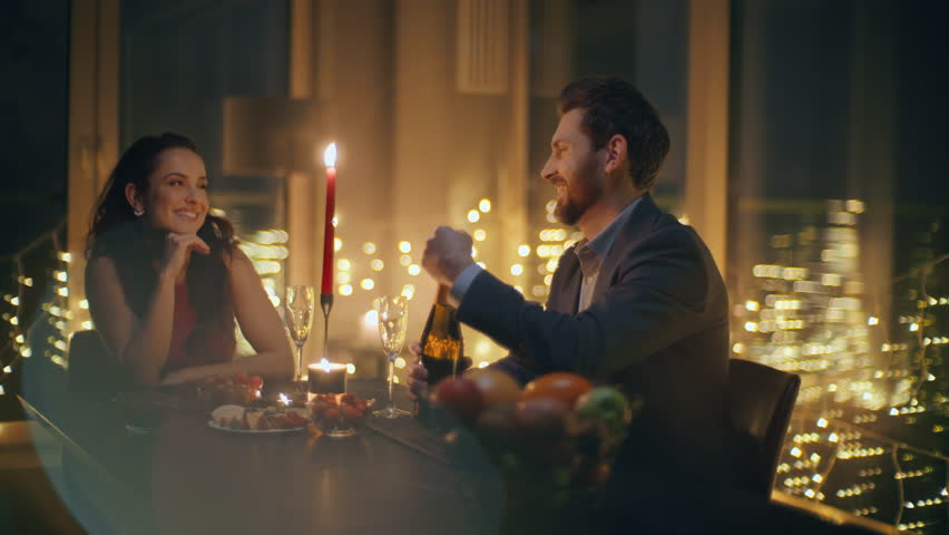 Enamoured pair celebrating anniversary at big windows hotel closeup. Happy gentleman opening champagne bottle pouring glasses at romantic date. Smiling woman holding glass enjoying moment with husband