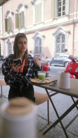 A lively young lady, sporting a checkered shirt, relishes her breakfast in the vibrant atmosphere of Bologna, Italy.