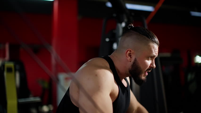 Muscular bearded man in black tank top performing cable fly exercise in gym, showcasing intense focus and effort
