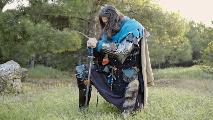 A man in a medieval knight costume stands kneeling and leaning on a sword stuck in the ground in front of him in the bright rays of the sunset. Close-up. Historical reconstruction