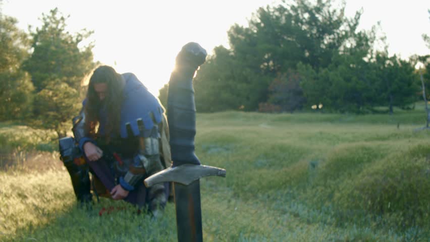 A man with a beard in a medieval knight costume stands kneeling in front of a sword stuck in the ground in the bright rays of the sunset. Pose of grief and thirst for revenge. Close-up