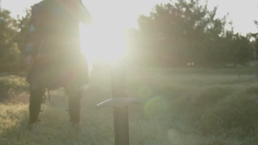 A man in a medieval knight costume approaches a sword stuck in the ground in the bright rays of sunset and takes it. Close-up. Historical reconstruction