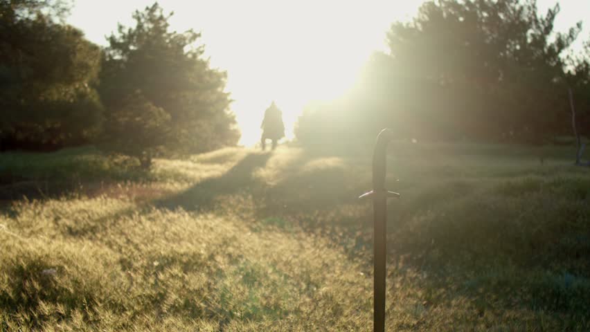 Full length portrait A man with a beard in a medieval knight costume slowly and carefully walks towards a sword stuck in the ground on the edge of a forest at sunset. Close up