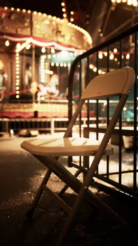 A white chair is placed on a balcony overlooking an abandoned carousel in a deserted amusement park at night. Multiple lights illuminate the darkness around old, unused rides.