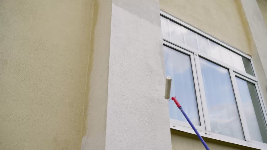 Painting the facade of a house with a long-handled roller. Worker holding brush, paint roller in yellow on house wall, crafts and renovation concept. Updating the appearance of the building.