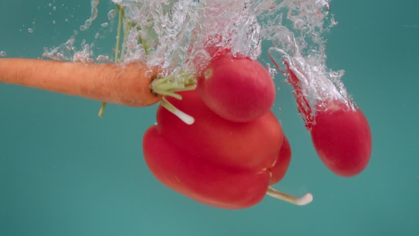 The vegetables of red pepper, green onions, carrot and tomatoes on a turquoise background fall and immerse in water, rotate, float and form bubbles. The concept of preparing food for cooking. Slow