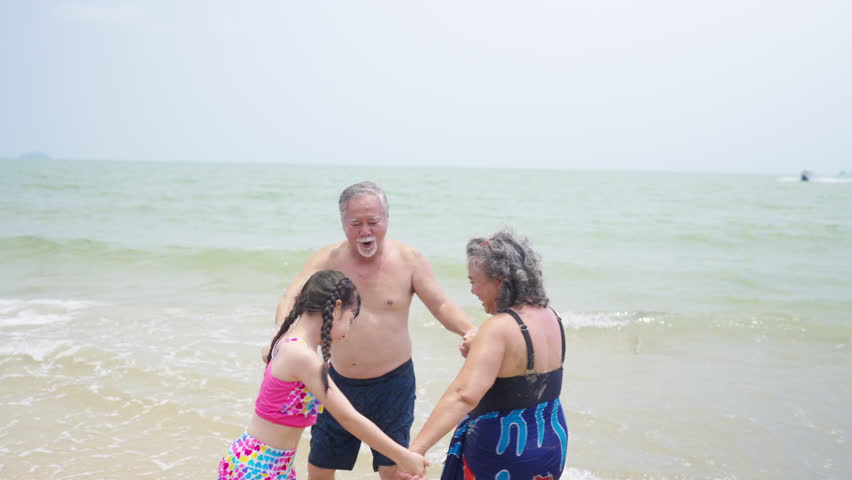 4K Happy Asian family enjoy and fun outdoor lifestyle travel ocean on summer holiday vacation. Grandparents and grandchild girl in swimwear playing sea water together at tropical beach in sunny day.