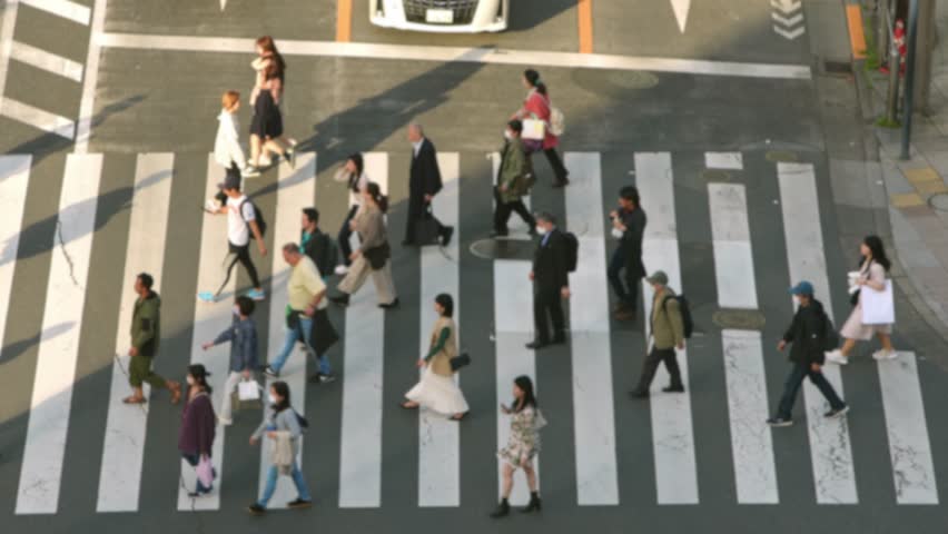 Out of focus shot of slow motion looking down on the intersection from a high vantage point, crowds of people hurry across the street in silhouette with cinematic shadows with sunset light.