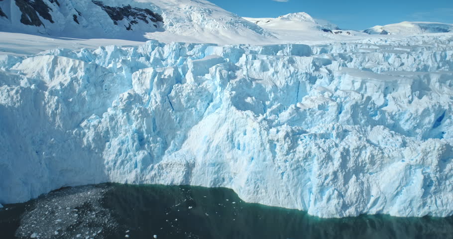 Zoom out panorama giant glacier ice wall iceberg towering on Antarctic coastline polar ocean. Sunny day blue sky, snow covered mountains. Ecology, global warming and climate change. Aerial drone shot