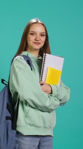 Vertical video Portrait of smiling woman with backpack holding school notebook and notes, isolated over studio background. Cheerful girl with school supplies in arms, preparing to go to university