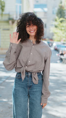 Hello. Caucasian young woman smiling friendly at camera, waving hands gesturing hi, greeting or goodbye, welcoming with hospitable expression outdoors. Lady girl on urban city street. Vertical