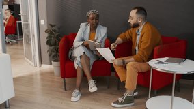 Full shot of young African American woman in ethnic turban, with tablet and Caucasian man in yellow knit cardie sitting in red armchairs in modern office, talking, planning marketing campaign - Powered by Shutterstock - Get 15% off with code: PIKWIZARD15