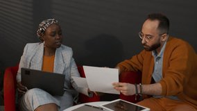 Medium shot of young African American female marketing specialist in bright ethnic turban and Caucasian male analyst sitting in red armchairs in office, studying data sheets and typing on laptop - Powered by Shutterstock - Get 15% off with code: PIKWIZARD15