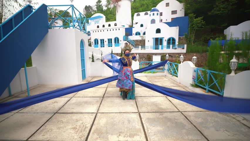 Person in vibrant traditional costume dancing in a picturesque setting resembling a Greek village, with white and blue buildings in the background.