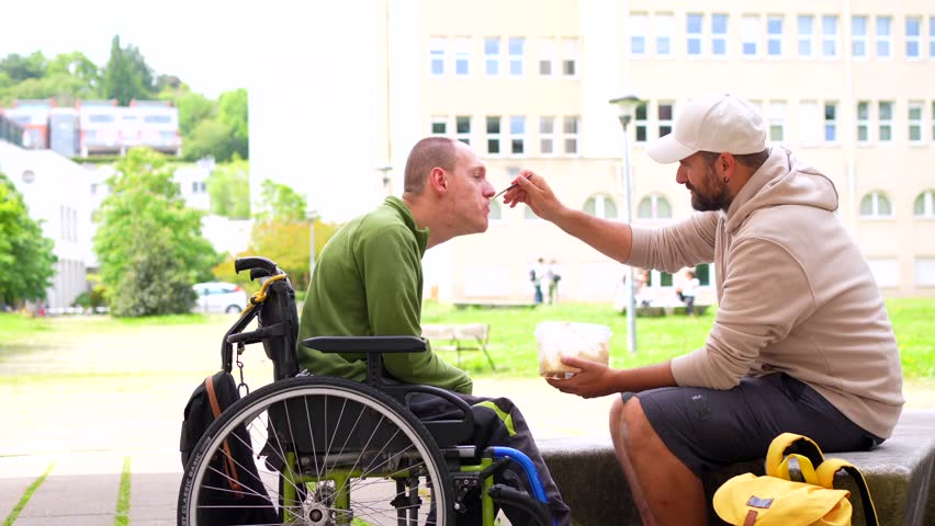 Full length photo of a caucasian adult college student feeding a friend with cerebral palsy on campus