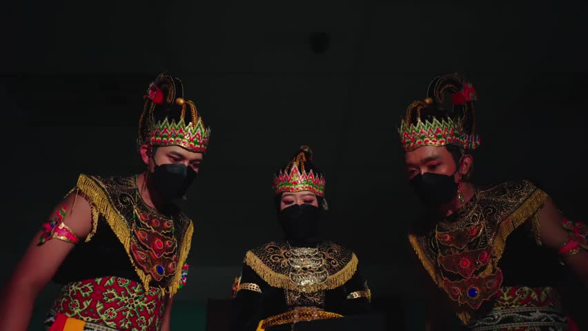 Three performers in traditional, ornate costumes and masks, illuminated by warm stage lighting, with a focus on cultural dance or theatrical presentation.
