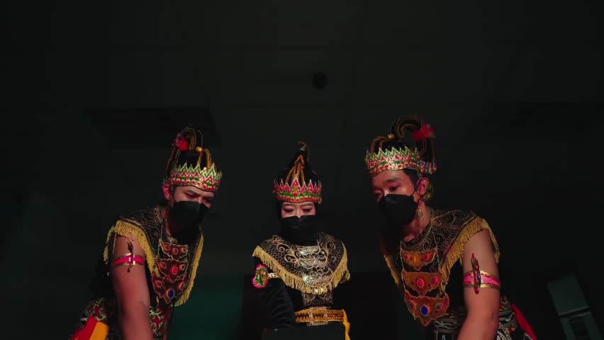 Three performers in traditional Javanese dance costumes and masks, with intricate headgear, captured in a dimly lit setting, showcasing cultural arts.