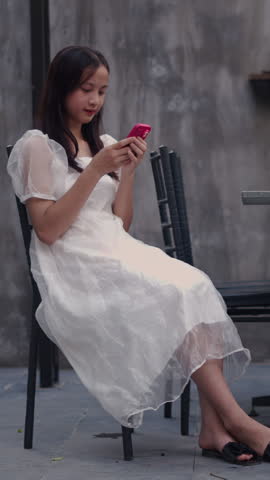 young asian teenager girl sitting on a terrace chair next to the table consulting her smartphone in summer