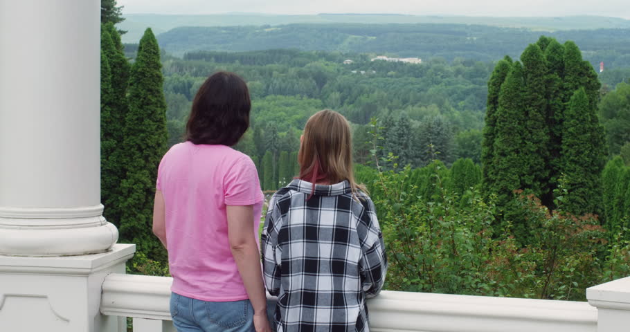 Family mom woman teen girl daughter admiring nature in landscaped park standing in gazebo, back view. Summer time with green trees in wide area. Walking together pastime leisure on weekend concept.