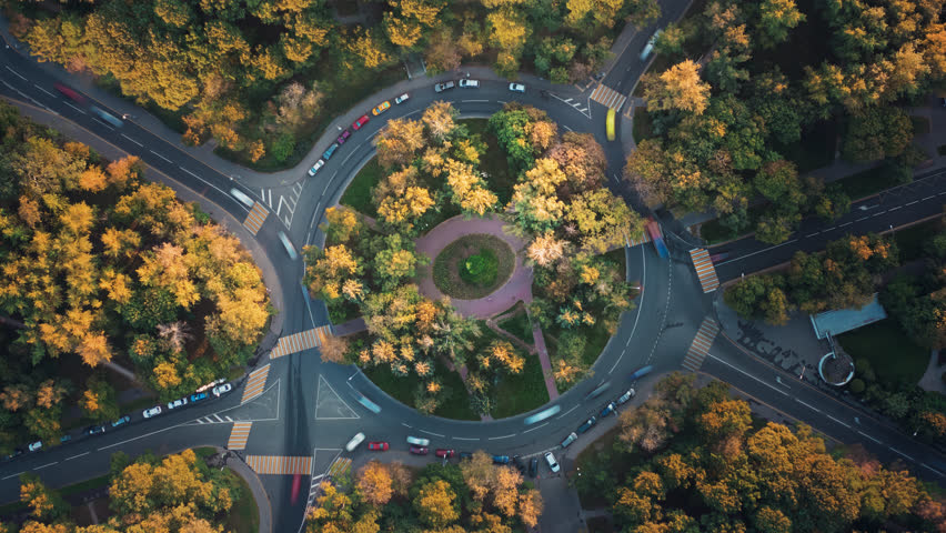 Top down time-lapse aerial view of a park with a roundabout intersection with slow camera rotation, beautiful sunset illuminates trees. Cars move fast along the road during an autumn traffic jam.