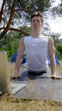Black-haired camper does workout on black mat looking in laptop. Young man finds online video course of yoga and decides to try exercises near camp tent against sky Vertical Shot.