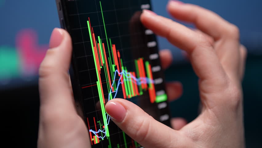Closeup of female stock trader holding smartphone with stock shares trading app on screen. Financial analysis of shares, stock shares exchange charts on display. Concept of investing money on share. - Powered by Shutterstock - Get 15% off with code: PIKWIZARD15