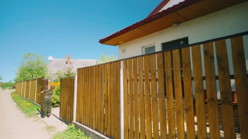Cute girl opening yellow wooden fence in front of house while blossom leaves fall down