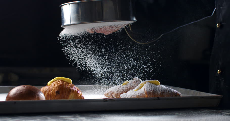 Super slow motion macro of professional artisan baker chef sprinkles sugar icing powder with sieve on homemade pastries sweet delicious dessert French croissants dessert in traditional bakery kitchen.