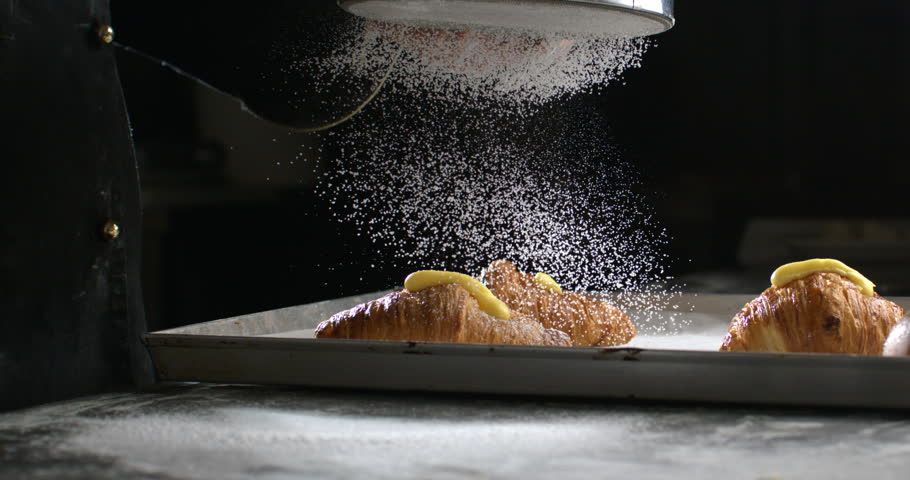 Super slow motion macro of professional artisan baker chef sprinkles sugar icing powder with sieve on homemade pastries sweet delicious dessert French croissants dessert in traditional bakery kitchen.