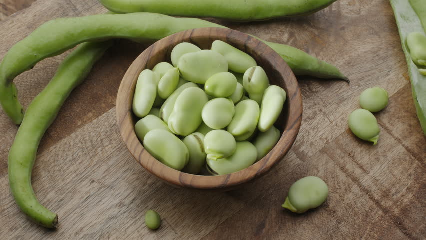 Broad beans in a wooden bowl and broad bean pods near it on a wooden table. Slow rotation of objects in frame video. 