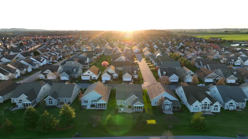 Suburban neighborhood at sunset, with rows of similar houses, green lawns, and a street leading directly into the glowing sun on the horizon. Aerial truck shot.