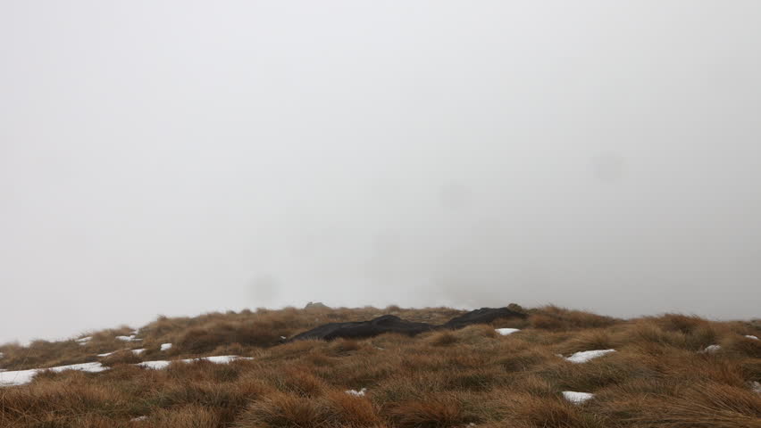 Time lapse of a man clad in an orange jacket pitches a tent in a snow-dusted mountainous area under a cloudy sky.