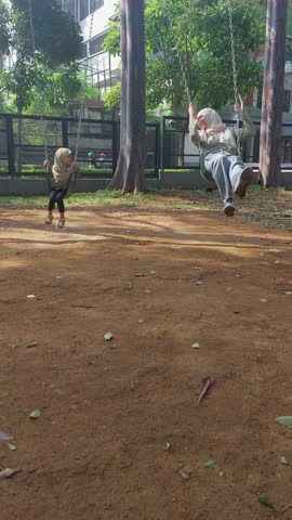Happy Asian mother and daughter in hijab smiling playing on swing together in park in summer, happy family concept