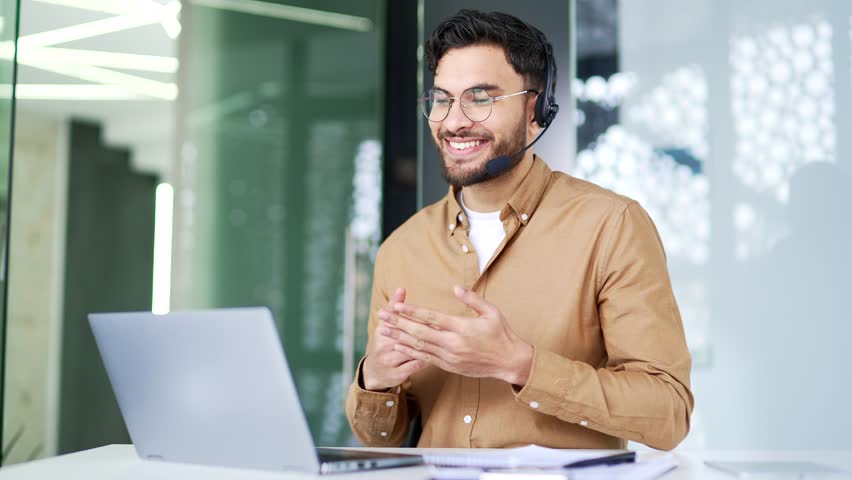 Confident manager talking on a video call using a laptop sitting at workplace in office. A businessman in wireless headset has a business meeting, speaks remotely, communicates at an online conference