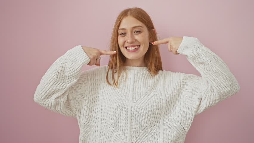 Radiant young redhead woman in a snug sweater, grinning and pointing at her healthy teeth, standing isolated over a joyful pink backdrop. dental care promo.