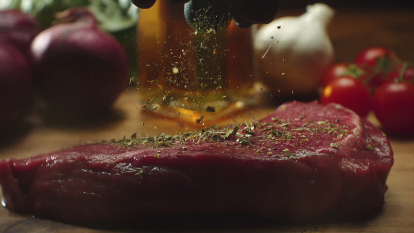 Chef hands sprinkling spices on an appetizing red raw steak on wooden chopped board. Preparing grilled tasty beef meat at restaurant kitchen on dark background. Cooking process in slow motion 1000fps