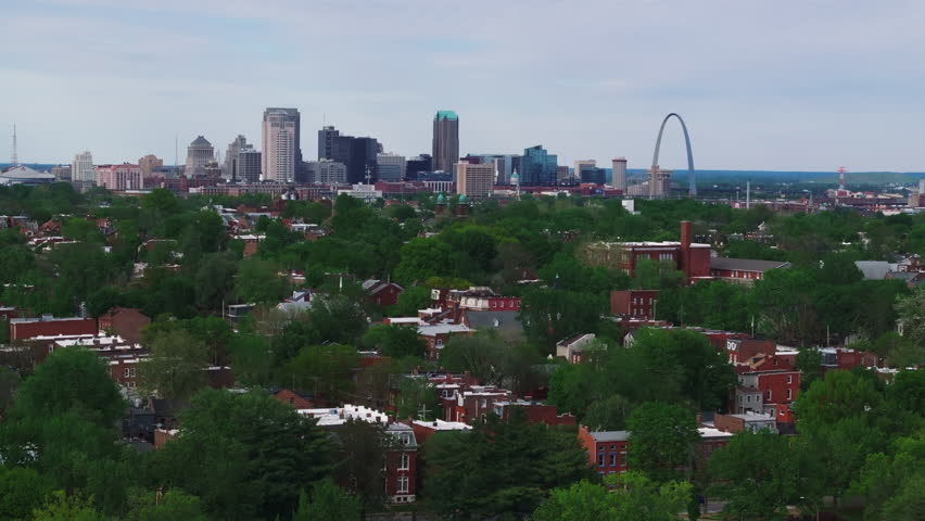 Aerial view of the St. Louis skyline, featuring the iconic gateway arch and lush greenery. Low buildings of a peaceful residential area in the foreground