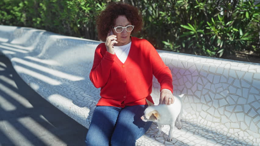 An elderly woman in a red sweater talks on the phone while sitting on a bench outdoors, accompanied by her white chihuahua in an urban park setting.