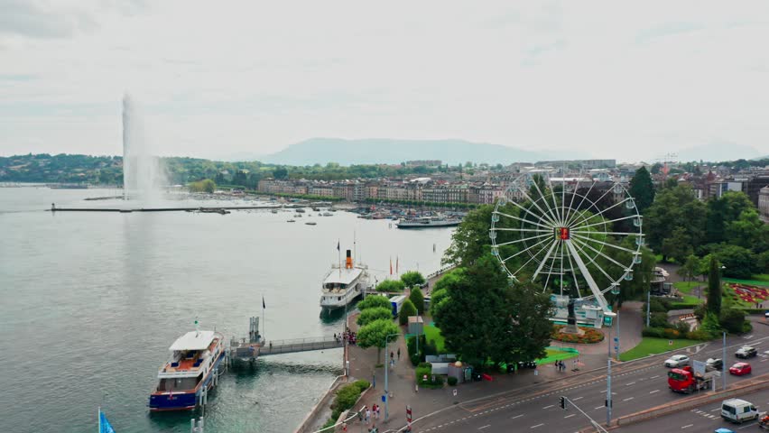 Beautiful aerial of Pont du Mont-Blanc bridge in Geneva, Switzerland. A large Ferris wheel and Jet d'Eau de Genève are seen while the drone flies backwards