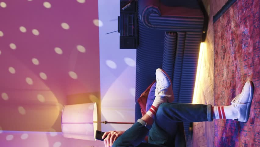 Portrait of young handsome creative man spending free time at retro disco party. Attractive guy sitting on soft chair wearing wireless headphones and listening to driving dance music.