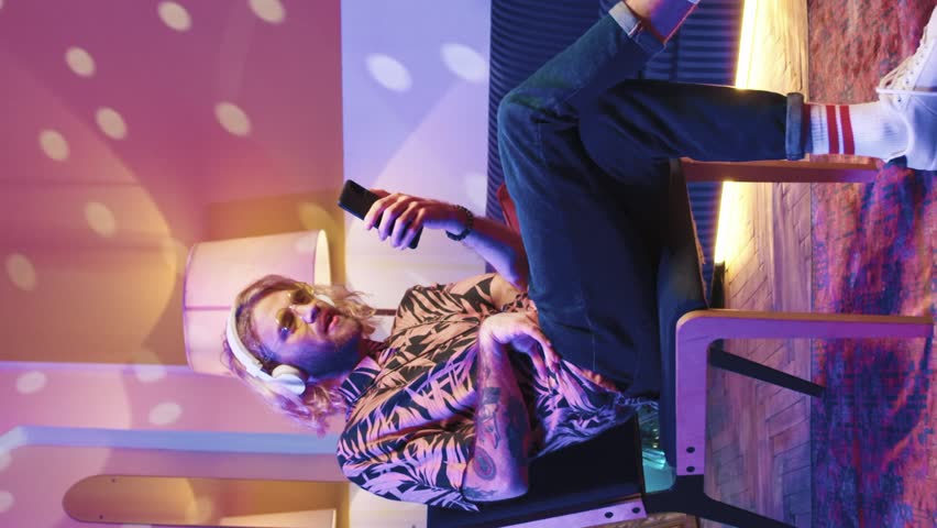 Attractive guy sitting on soft chair wearing wireless headphones and listening to driving dance music, spending time in social media and internet, chatting, texting by phone. Having fun at home party.